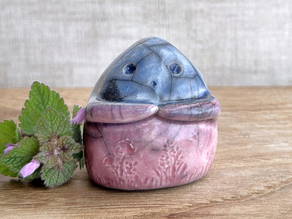 a vaguely dome-shaped nature spirit ceramic sculpture. it is glazed in a bottom-to-top gradient of mauve to blue and has plant and flower carvings on its body. it also has a gentle face, and two little arms across its front. there are tiny dark grey spiderweb patterns in the glaze, in addition to the normal raku crackles, from where raindrops fell on it.