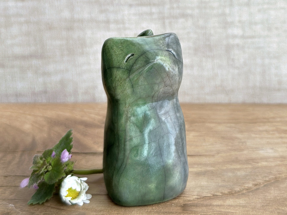 Raku ceramic earth and water spirit kitsune sitting fox sculpture glazed in green and grey. It is sitting up with its tail curled up its back, and it has a gentle, kind face. 