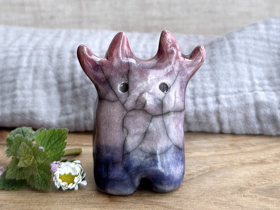 a raku ceramic creature. it has four stubby legs and a wide lower half. it tapers up a little and eventually ends in four small antlers. on the way, it has a very sweet face with a cute little nose. it's glaze in a bottom-to-top gradient of dark blue to purple to a soft red purple for the antlers. it is a friend.