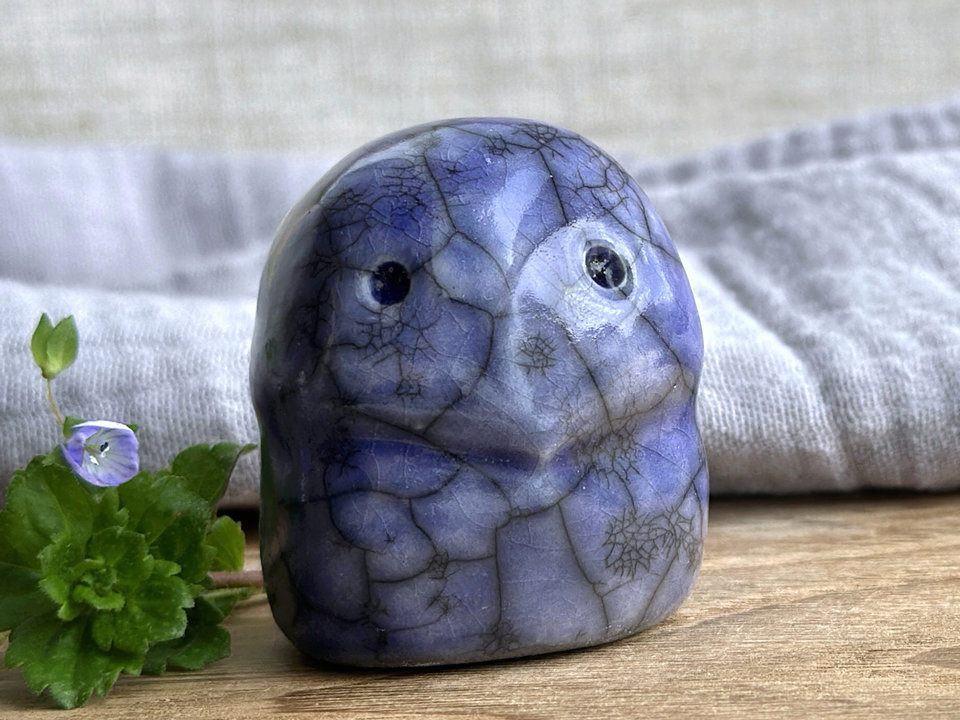 A kodama nature and forest spirit kodama kami ceramic raku sculpture glazed in a deep indigo-violet colour. It has a gentle face.  There are tiny dark grey spiderweb patterns in the glaze, in addition to the normal raku crackles, from where raindrops fell on it.