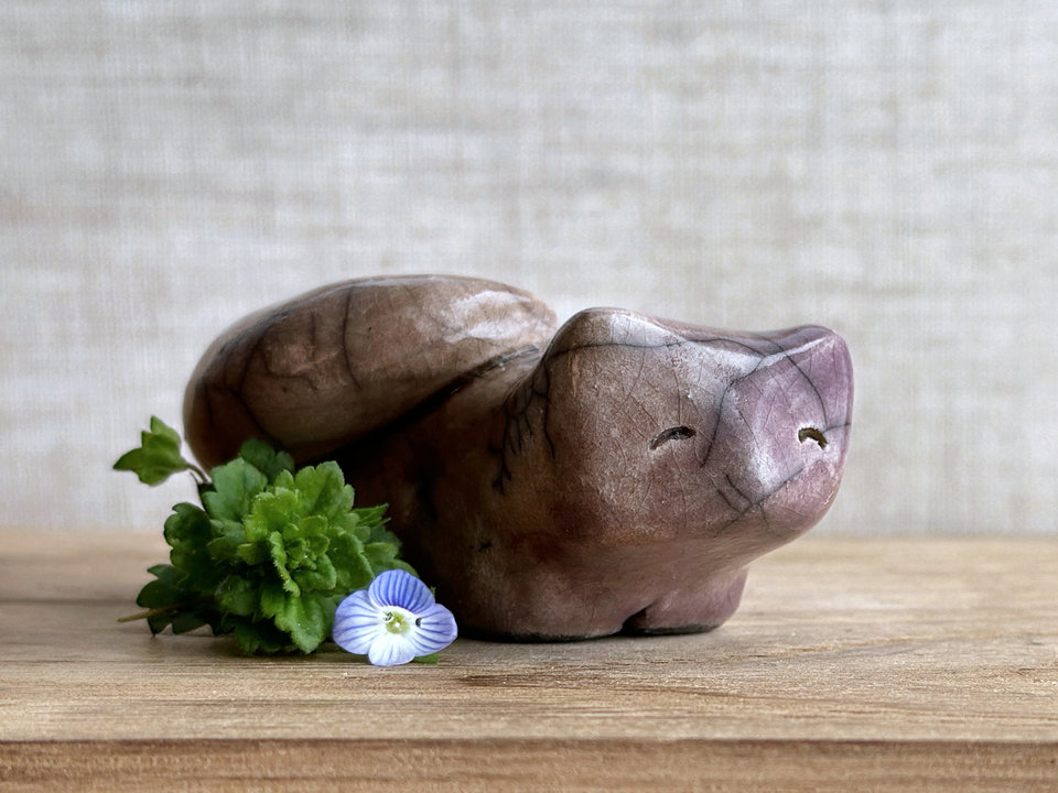 raku ceramic kitsune fox sculpture glazed mostly on its right side in soft brown and mostly on its left side in purple. it has a kind, gentle face, and a tail curled over its back on its right side.