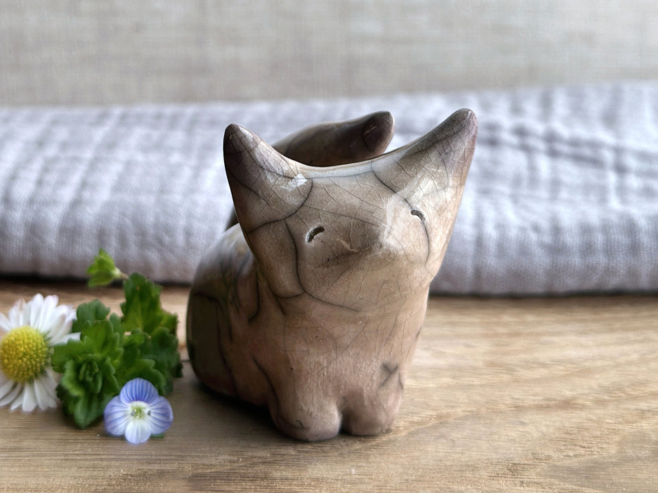 Sitting rake ceramic kitsune fox sculpture glazed in pale brown with soft violet tips to its ears and tail. It has a kind, gentle face, and its tail is curled up its back. It has two little feet at the front and rather large, pointy ears.