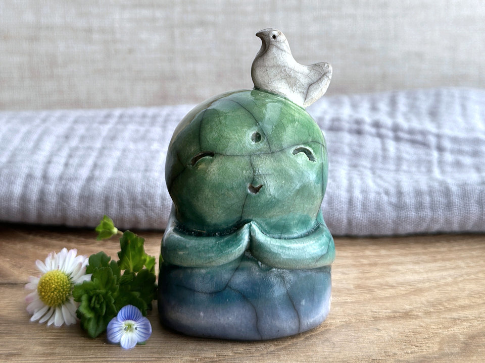 a shinto jizo nature spirit guardian raku ceramic sculpture glazed in a bottom-to-top gradient of blue to forest green to spring green. they have a happy, smiley face, little praying hands, and a sweet white bird on their head.