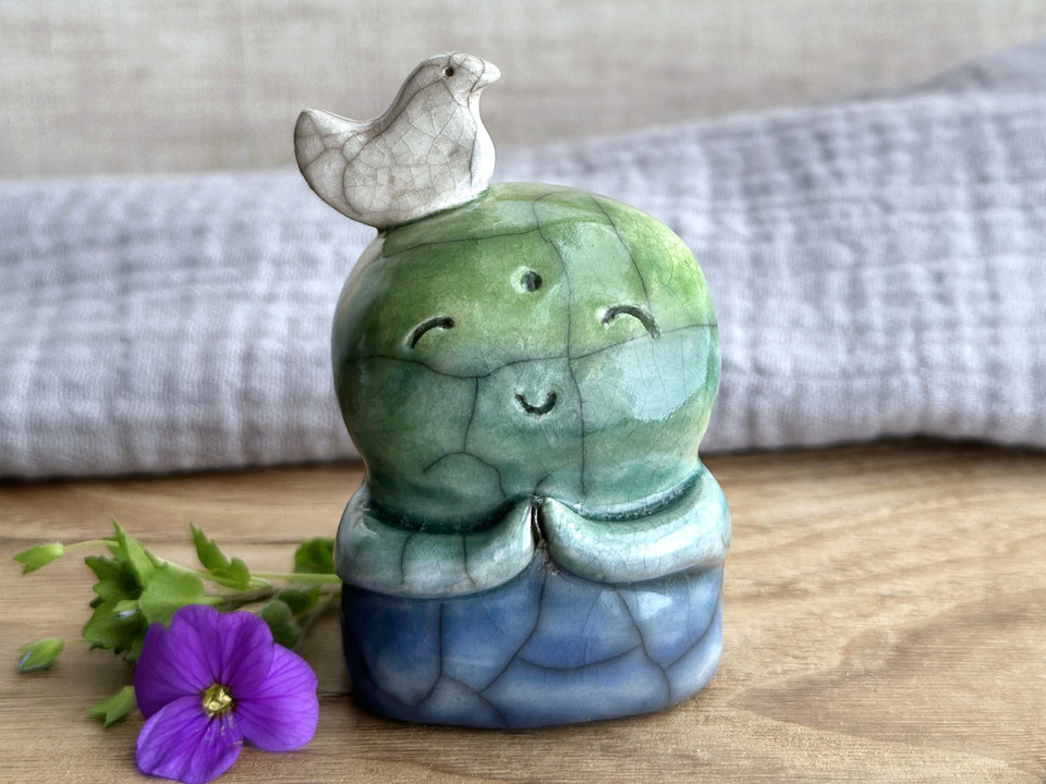 rweb patterns in the glaze, in addition to the normal raku crackles, from where raindrops fell on it.a raku ceramic jizo nature spirit guardian sculpture glazed in a bottom-to-top gradient of blue to forest green to spring green. they have a happy, smiley face, little praying hands, and a sweet white bird on their head.