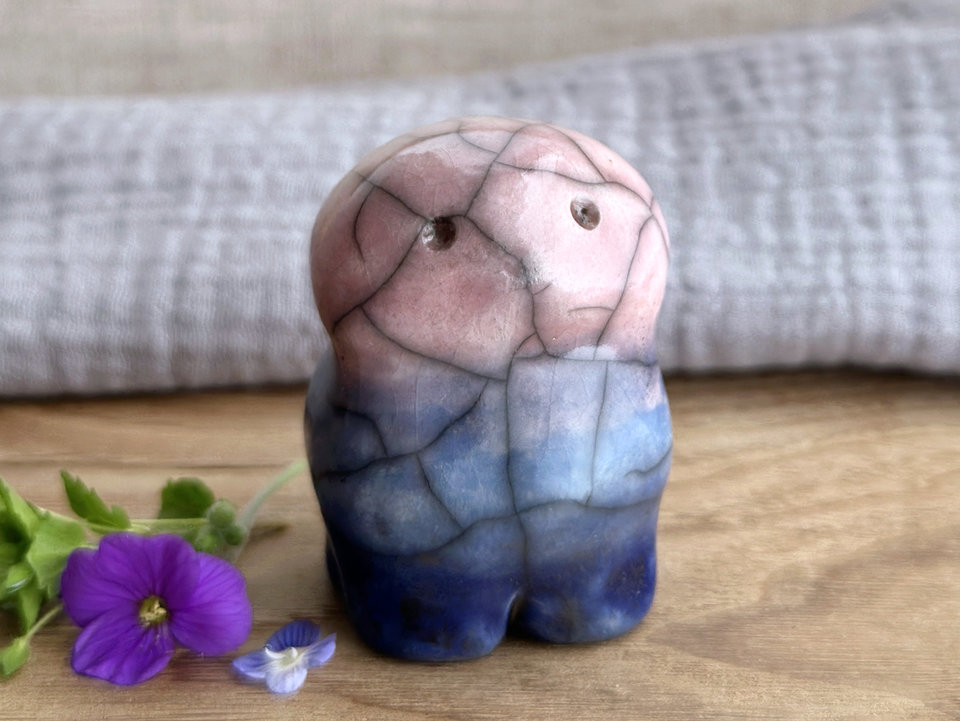 a raku ceramic creature. it has four stubby legs, a widish lower half, and a round head with a small nose and two round eyes. it tapers up a little and eventually ends in four small antlers. on the way, it has a very sweet face with a cute little nose. it's glaze in a bottom-to-top gradient of dark blue to blue to a pale pinky red. it is a friend.