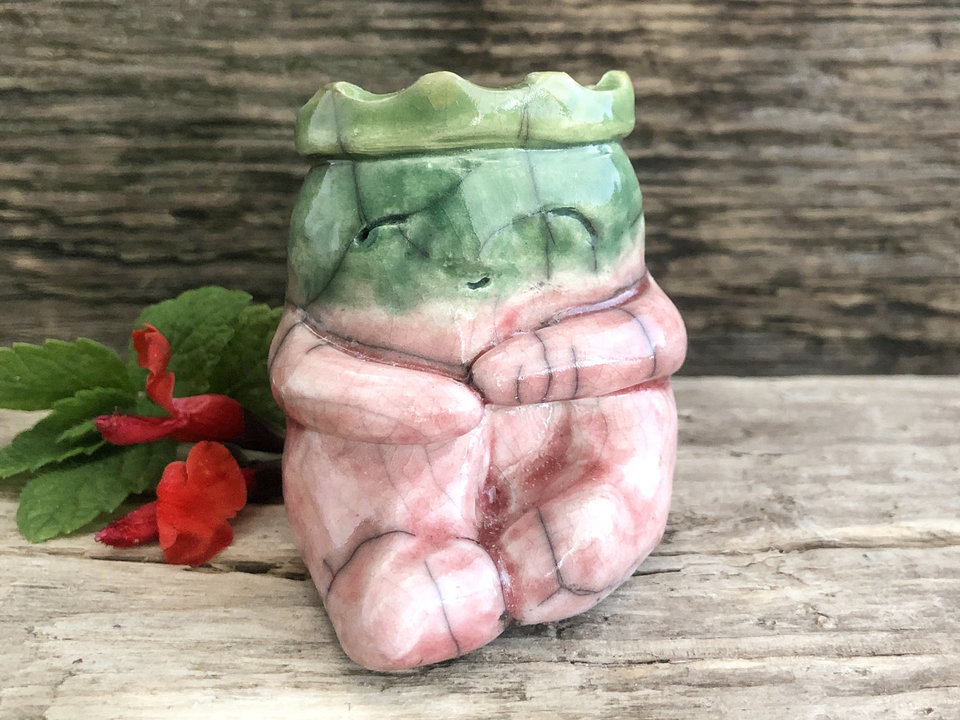 Raku sculpture of a cute sitting creature glazed in soft pink and greens with a green and golden yellow crown on his head and a very happy face.