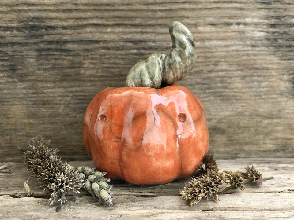 Raku ceramic pumpkin glazed in warm red-orange. It has two eyes, a serious expression, and a grey-green-brown twisty stem.