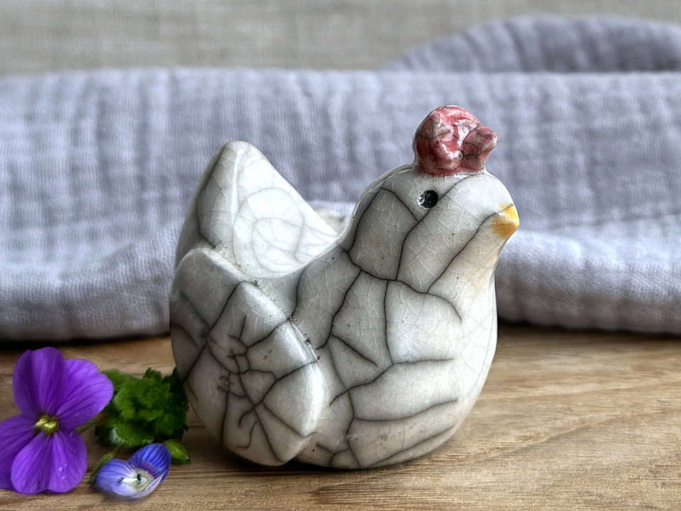 a white raku ceramic chicken with grey eyes, a pale orange beak, and a bordeaux red comb on the top of her head. she looks very sweet.