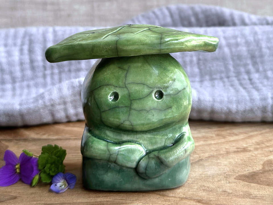 Raku ceramic sculpture glazed in forest green to spring green (bottom to top) with a kind, but slightly intense, face, and a brighter yellow-green leaf on its head. It also has two cute little arms.