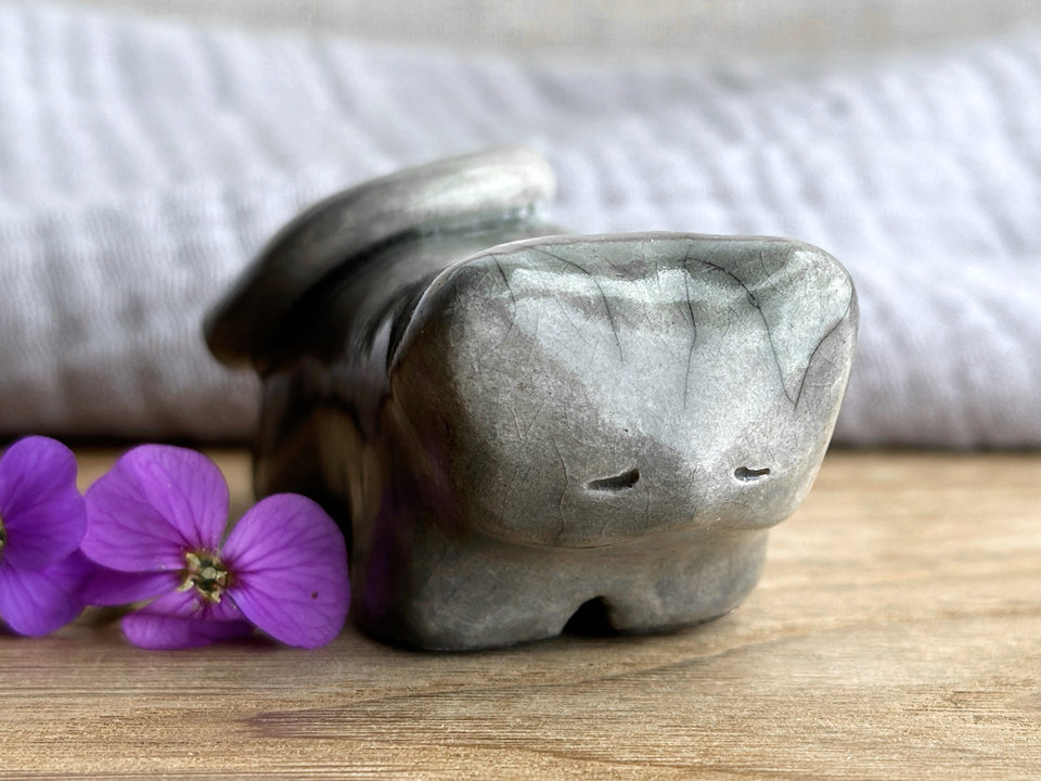 Little raku ceramic cat spirit kami sculpture glazed in grey. It has a kind, gentle, sleepy face, and its tail is curled over its back on its right.