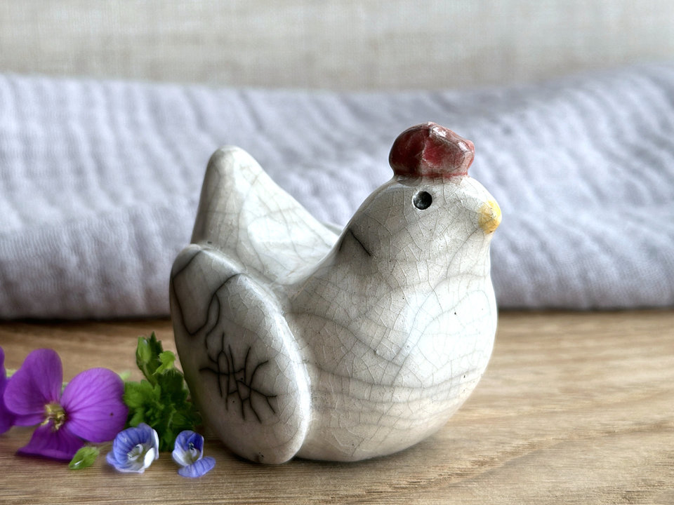 a white raku ceramic chicken with grey eyes, a pale orange beak, and a bordeaux red comb on the top of her head. she looks very sweet.