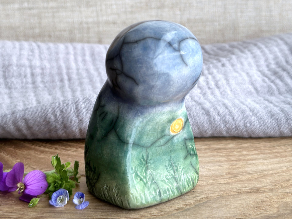 Raku ceramic earth spirit sculpture glazed in spring green, forest green, and blue (bottom to top). It has plant shapes and flowers carved into her and a yellow sun carving too.