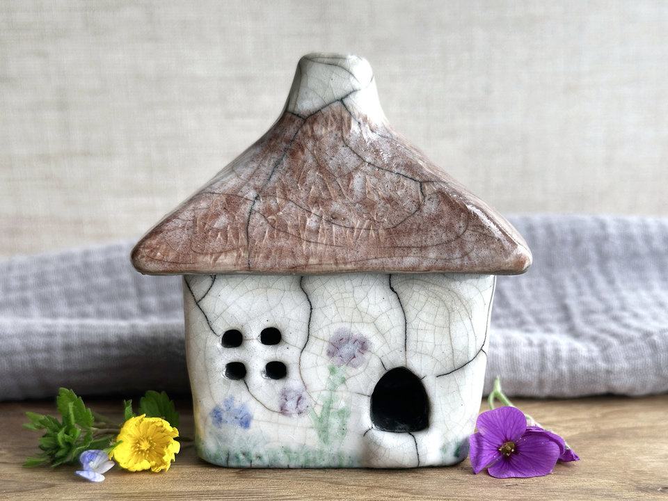 a rustic raku ceramic kurinuki cottage glazed in white with green plant carvings and purple, violet, and indigo flower carvings on the walls. the roof has a chimney and is glazed in brown with hints of white and carved to look like thatch. the cottage has a cute little doorway and lots of windows.