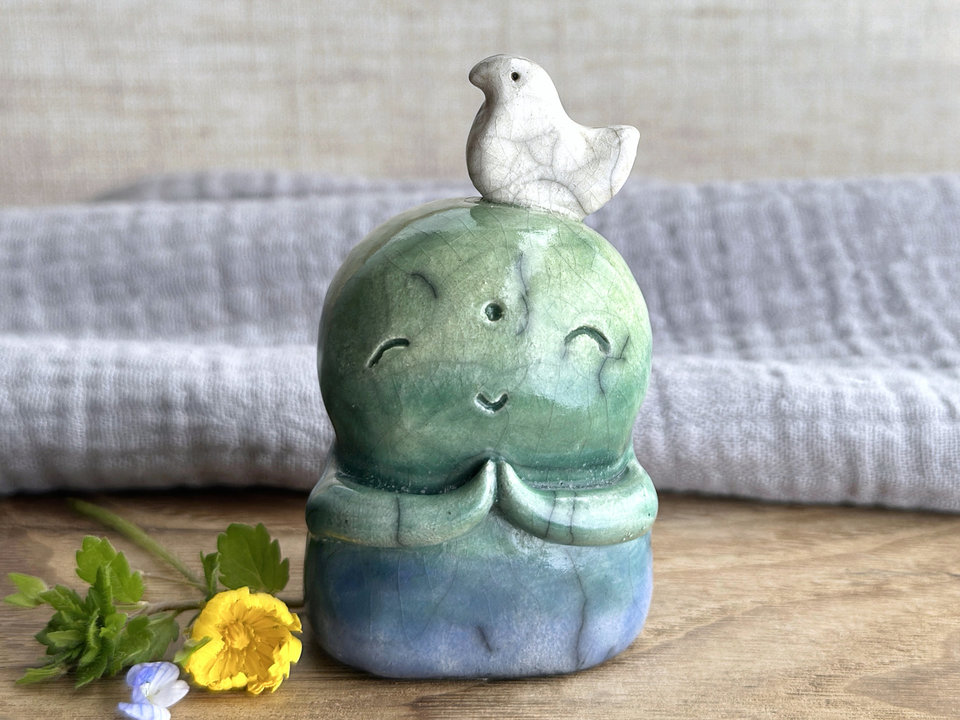 a raku ceramic jizo nature spirit guardian sculpture glazed in a bottom-to-top gradient of blue to forest green to spring green. they have a happy, smiley face, little praying hands, and a sweet white bird on their head.
