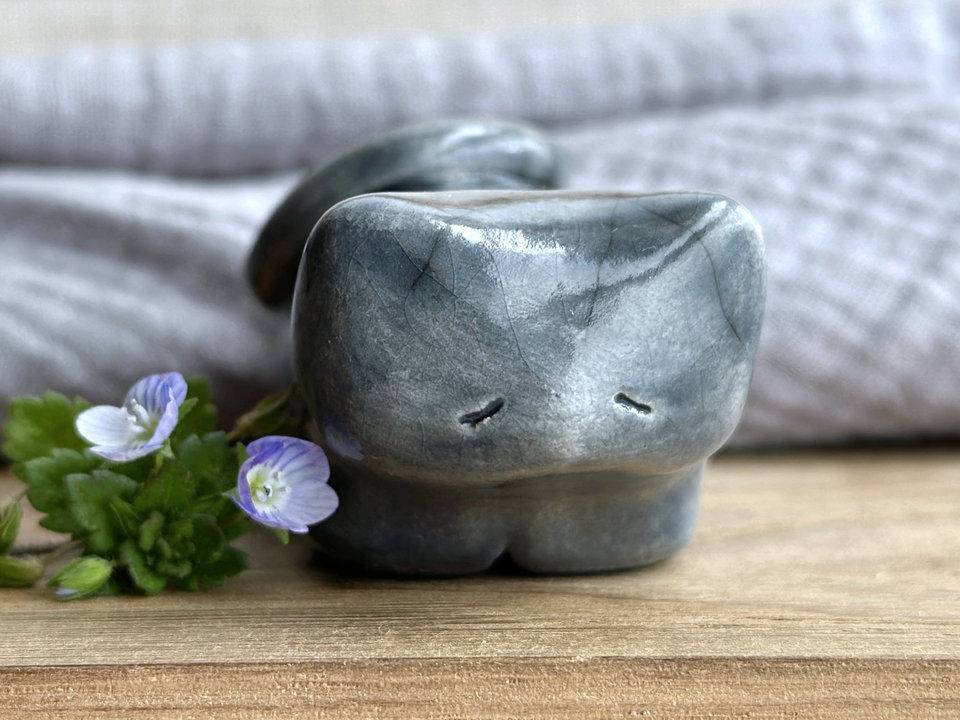 Little raku ceramic cat spirit kami sculpture glazed in grey. It has a kind, gentle, sleepy face, and its tail is curled over its back on its right.