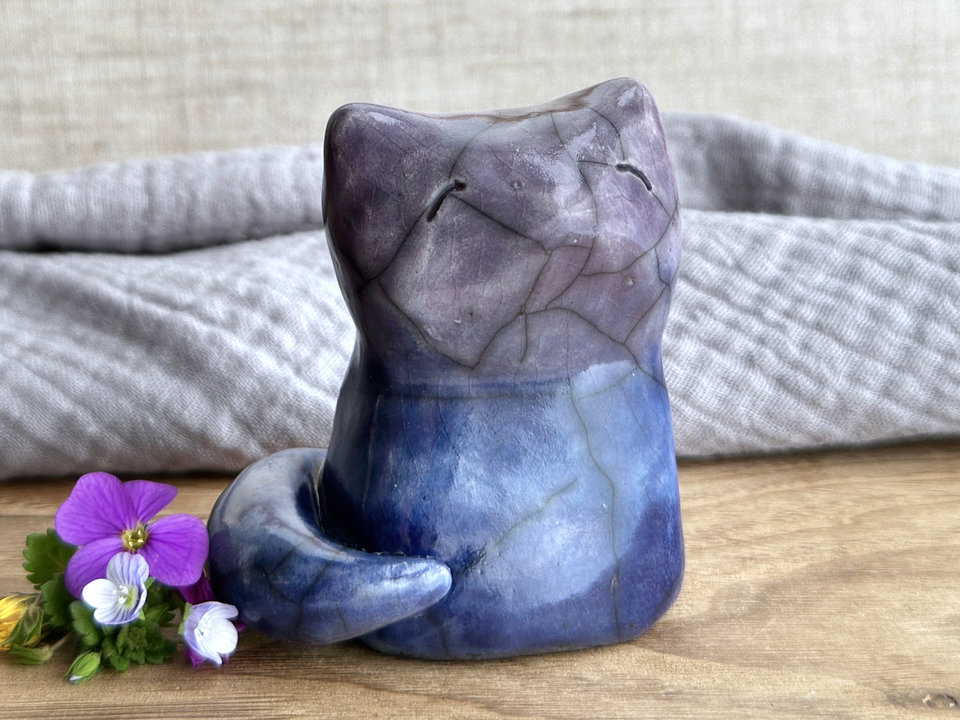 A calm and gentle Shinto Inari nighttime kitsune fox spirit ceramic raku statue glazed in a bottom-to-top gradient of dark blue to blue to purple. Its tail is curled around the right side of its body, and it has a kind face. There's a mild glaze disturbance pattern on its face that look like a little bit like where tiny snowflakes fell.