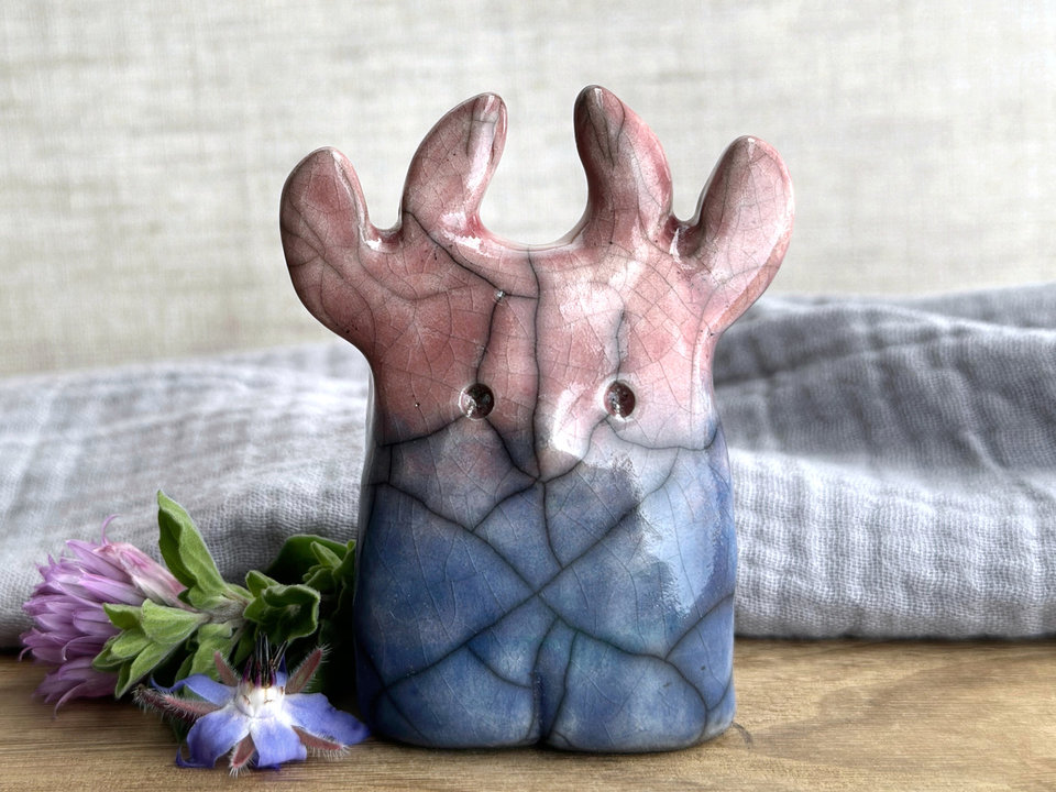 a raku ceramic creature. it has four stubby legs and a wide lower half. it tapers up a little and eventually ends in four small antlers. on the way, it has a very sweet face with a cute little nose. it's glaze in a bottom-to-top gradient of blue to soft reddish pink. it is a friend.