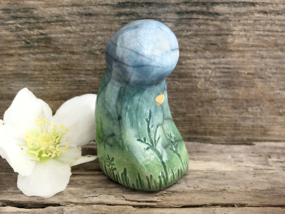 Raku ceramic mother earth spirit spirit sculpture glazed in spring green, forest green, and blue (bottom to top). It has plant shapes and flowers carved into it and a yellow sun carving too.