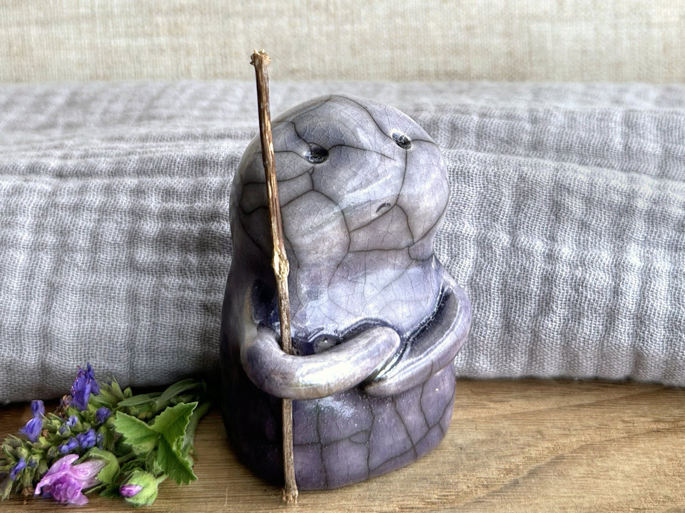 raku ceramic nature spirit warrior sculpture. it is glazed in a bottom-to-top gradient of violet to pale violet and carries a brown stick in one of its arms. it has a sad and worried but determined look on its gentle face.