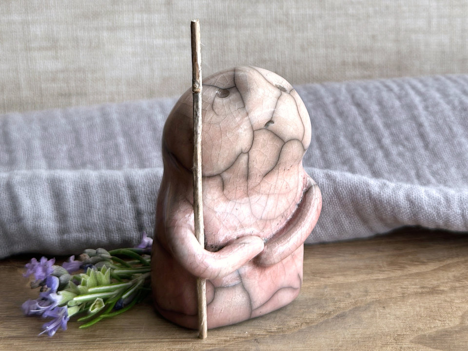raku ceramic nature spirit warrior sculpture. it is glazed in a bottom-to-top gradient of pale soft red to pale peach and carries a brown stick in one of its arms. it has a sad and worried but determined look on its gentle face.