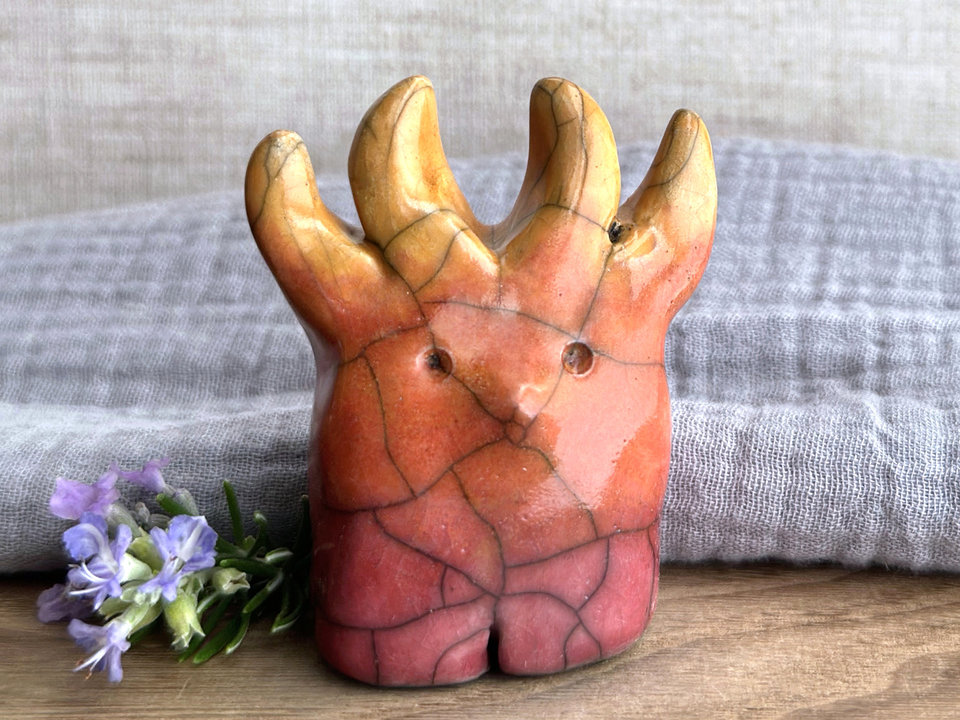 a raku ceramic creature. it has four stubby legs and gets wider as its body goes up. it eventually ends in four small antlers. on the way, it has a gentle face with a cute little nose. it's glazed in a bottom-to-top gradient of red-pink to warm orange to golden yellow for the antlers. it is a friend.