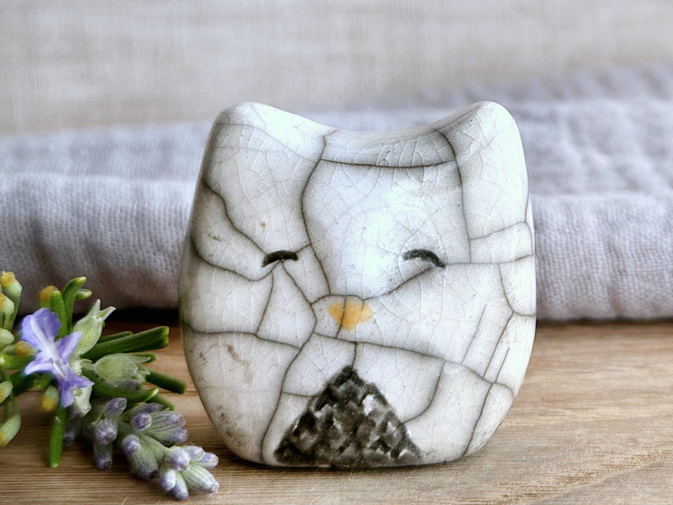 Raku ceramic little owl or owlet kami nature spirit statue with sort of pointed ears. It’s glazed in white and grey-brown with gentle, smiling eyes and a little golden yellow beak.