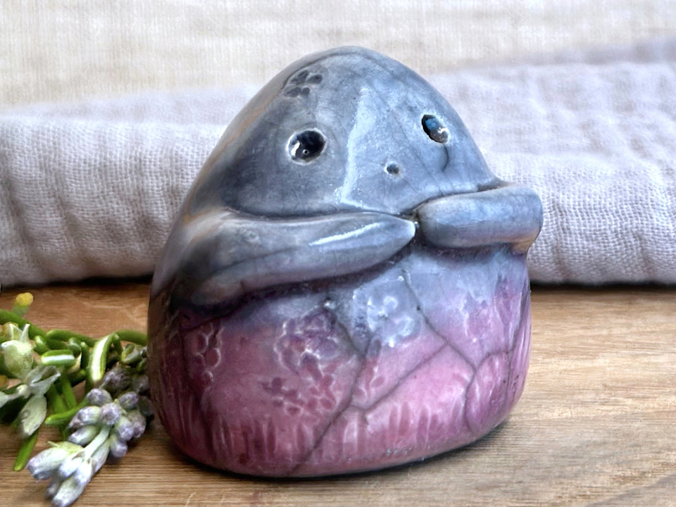 a vaguely dome-shaped nature spirit ceramic sculpture. it is glazed in a bottom-to-top gradient of soft mauve to blue and has plant and flower carvings on its body. it also has a gentle face, and two little arms across its front.