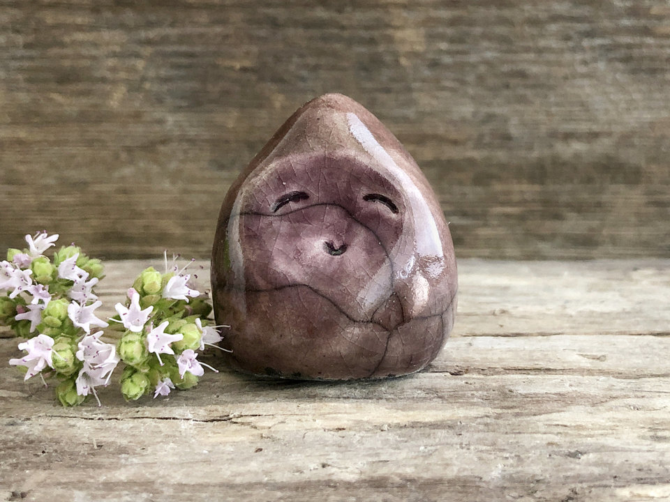 A small, almost (but not quite) teardrop-shaped raki ceramic earth spirit sculpture. It has a gentle face and is glazed in soft purple and warm brown colours.