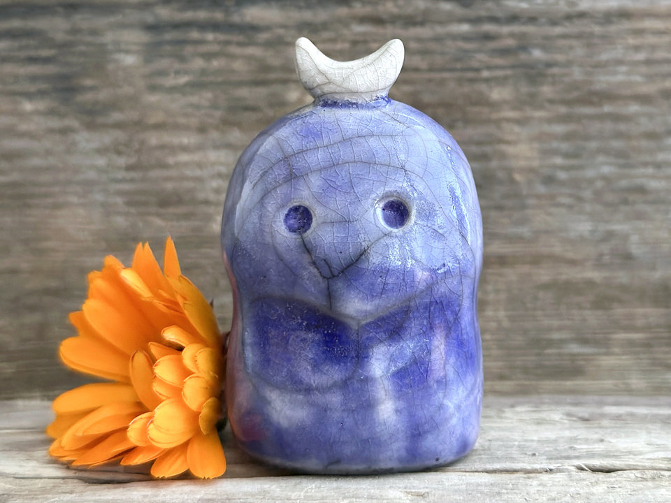 A kodama nature and forest spirit kami ceramic raku sculpture glazed in deep blue. It has a gentle face, and a white crescent moon on its head.