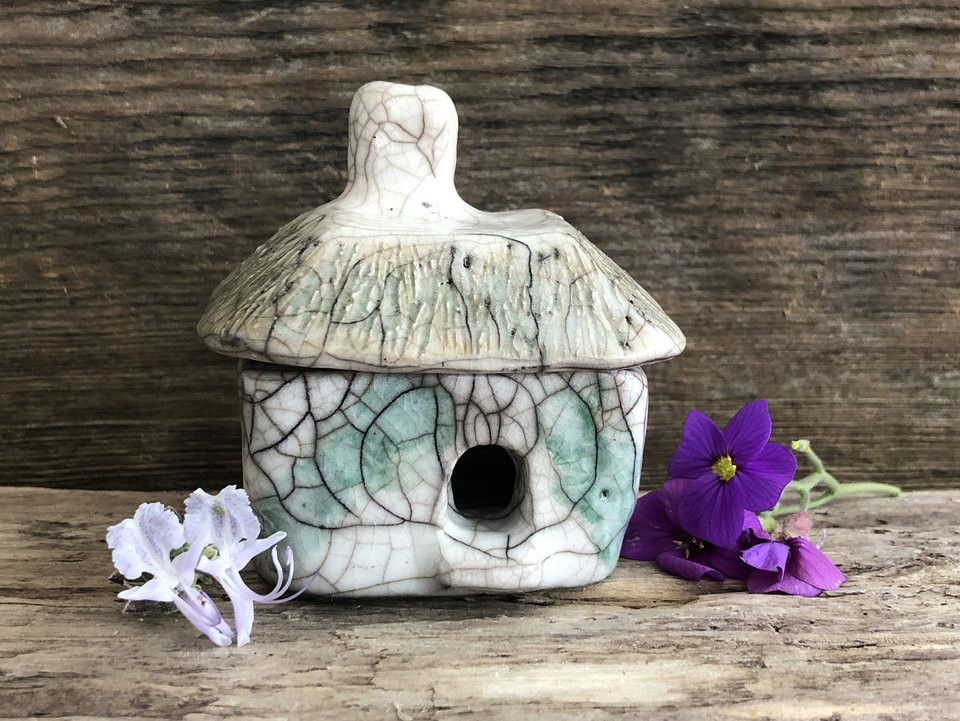 Raku ceramic kurinuki cottage glazed in white with green plant patterns on the walls. The roof has a chimney and is glazed in white, pale green, and light brown to look like thatch. It has a cute little doorway and several windows.