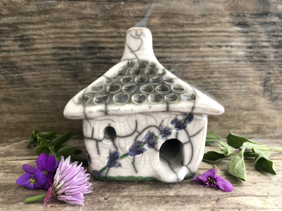 Raku ceramic kurinuki cottage glazed in white with green and purple lavender flower patterns on the walls. The roof has a chimney and is glazed in white, pale green, and light brown to look like thatch. It has a cute little doorway and lots of windows.