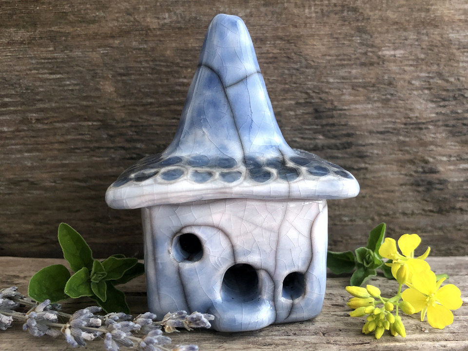 Raku ceramic kurinuki cottage glazed in blue and white with with a tall, pointed chimney. The roof below the chimney is glazed in white, grey, and blue to look like stone tiles. The house has a cute little doorway and lots of windows.