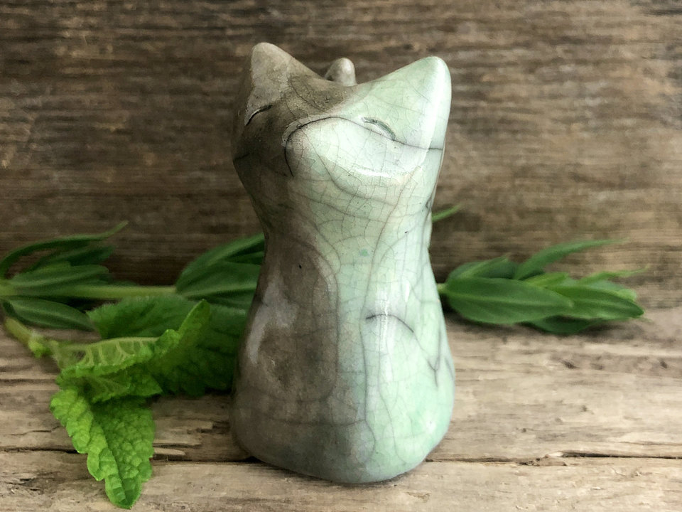 A happy Shinto Inari earth and water elemental spirit kitsune fox kami ceramic raku statue glazed in green, blue, grey, and brown. She is sitting up with her tail curled up her back, and she has a gentle, kind face. 