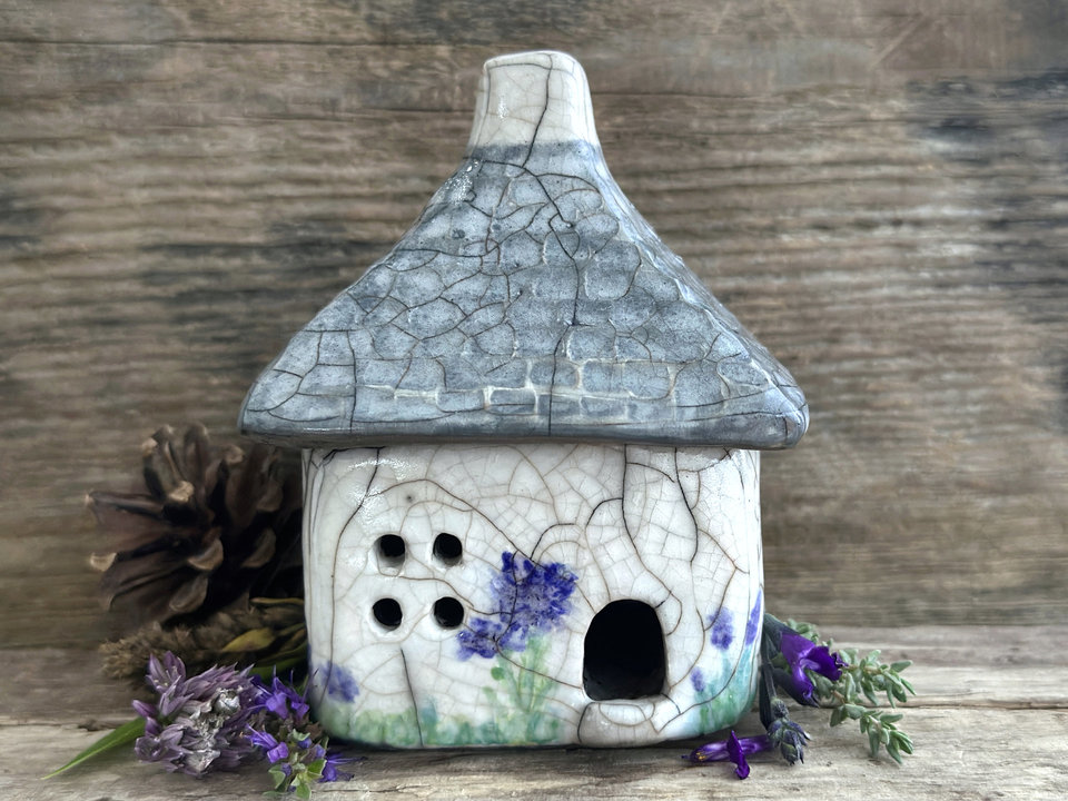 Rustic raku ceramic kurinuki cottage glazed in white with green plant and purple, violet, and indigo flower patterns on the walls. The roof has a chimney and is glazed in grey with hints of white to look like slate. It has a cute little doorway and lots of windows.