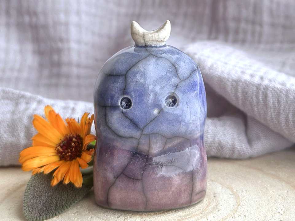 A kodama nature and forest spirit kami ceramic raku sculpture glazed in a bottom-to-top gradient of purple to blue. It has a gentle face, and a white crescent moon on its head.