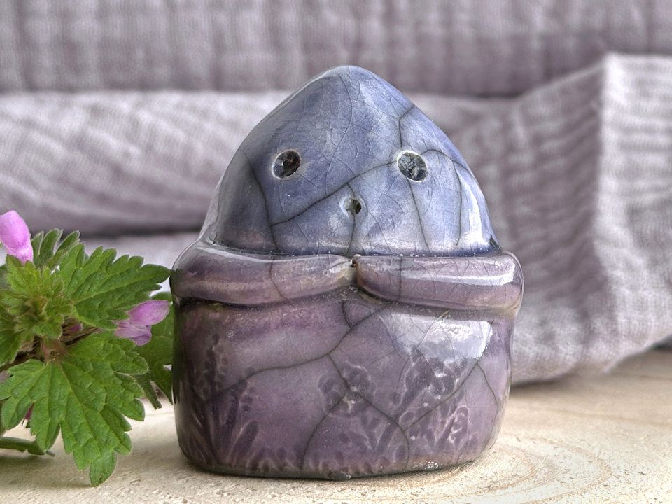 a vaguely dome-shaped nature spirit ceramic sculpture. it is glazed in a bottom-to-top gradient of violet to blue and has plant and flower carvings on its body. it also has a gentle face, and two little arms across its front.
