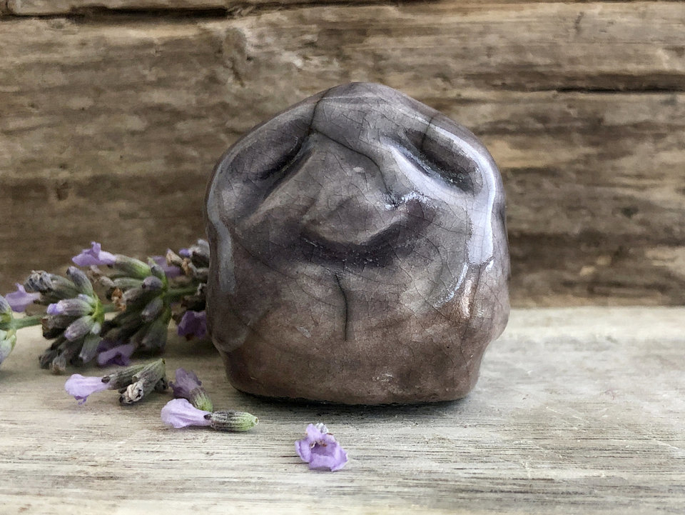 Raku ceramic sculpture of an elemental stone spirit gnome. He is glazed in soft violet and grey-brown and looks very natural, almost like his face was formed by weather and geology.