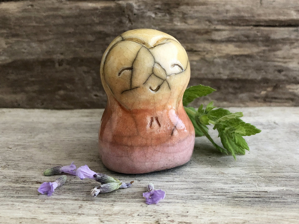 A smiley, loving, happy little Jizo Shinto raku ceramic sculpture talisman in shades of soft pink, orange, and yellow (from bottom to top) with a little heart around her face.