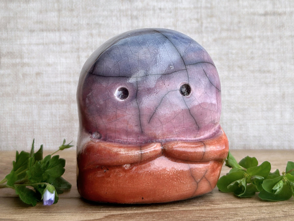 A kodama nature spirit ceramic guardian statue glazed in a top-to-bottom gradient of blue to violet to mauve to red-orange. It has a wide face, two big eyes, and cute little arms.