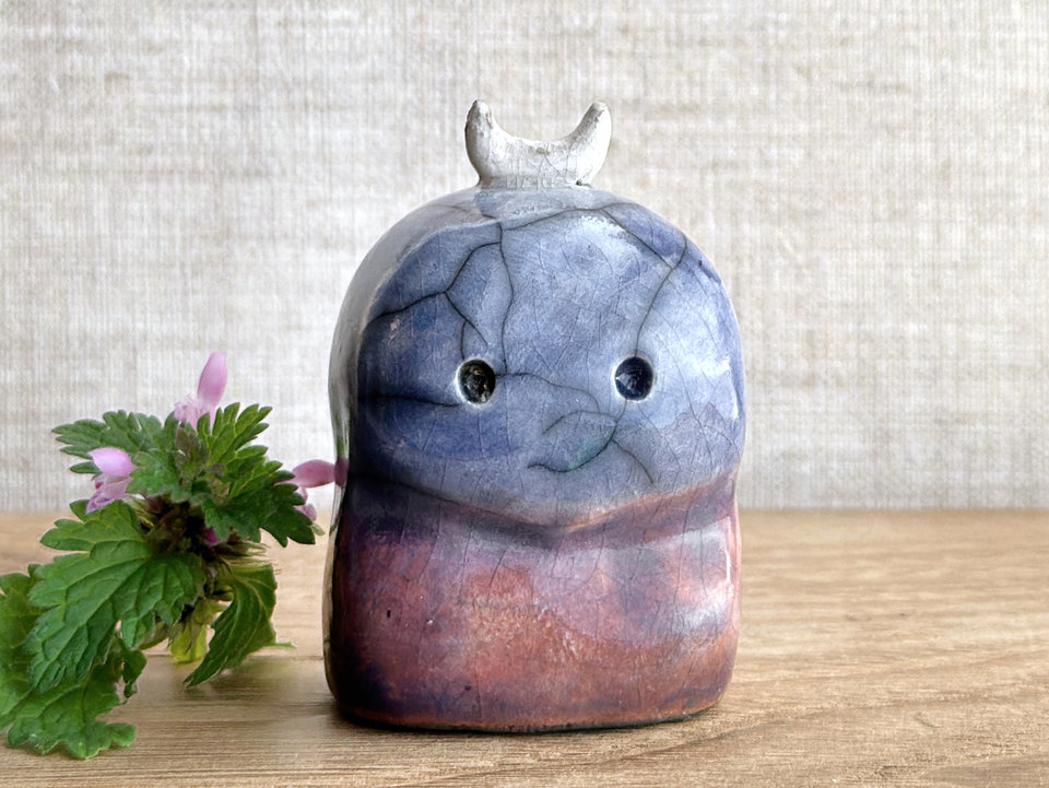 A kodama nature and forest spirit kodama kami ceramic raku sculpture glazed in a bottom-to-top gradient of red-orange to mauve to purple to indigo. It has a gentle face, and a white crescent moon on its head.