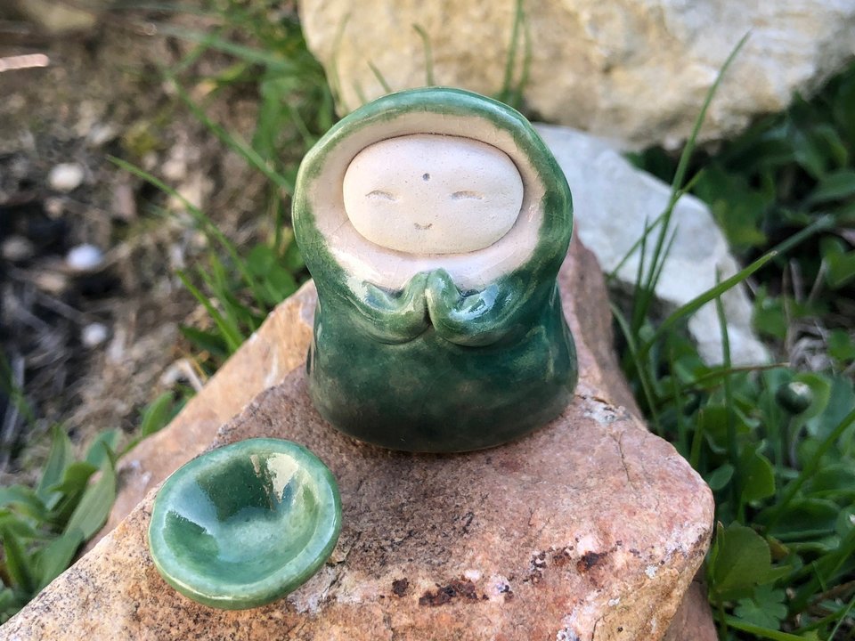 Ceramic sculpture of a little praying forest monk. His sweet face is unglazed white and he wears a green winter robe with a big hood. He has little light green mittens on his hands and a bowl to match.