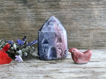 Tiny raku ceramic cottage glazed in indigo, violet, purple, and deep red, with a little red bird sat next to it. Its roof is carved to look like slates. It has a carved door and windows and lots of plant and flower carvings too.