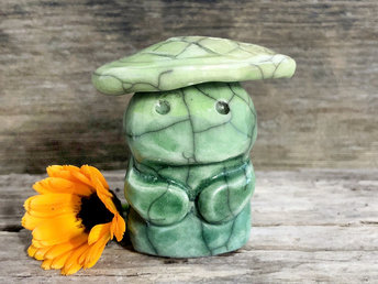 Raku ceramic sculpture glazed in forest green to spring green (bottom to top) with a sweet face and a lighter, yellow-green leaf on its head. It also has two cute little arms.