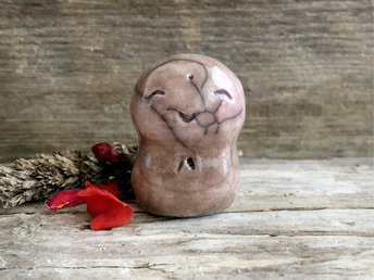 A smiley, loving, happy little Jizo Shinto raku ceramic sculpture talisman glazed in a bottom-to-top gradient of pale warm brown to soft pale brown-pink. It has a sweet, gentle face.