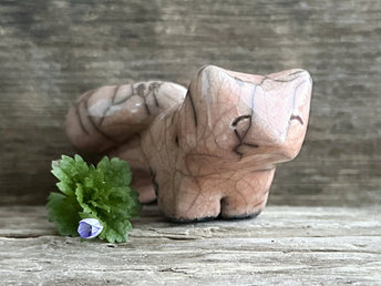 Raku ceramic kitsune fox sculpture glazed in a soft warm peach colour. It has a kind, gentle face, and a tail curled over its back. There are tiny dark grey spiderweb patterns in the glaze, in addition to the normal raku crackles, from where raindrops fell on it.