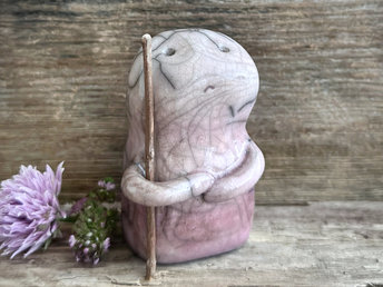 raku ceramic nature spirit warrior sculpture. it is glazed in a gradient from darker to lighter soft mauve plum (bottom to top) and carries a brown stick in one of its arms. it has a sad and worried but determined look on its gentle face.