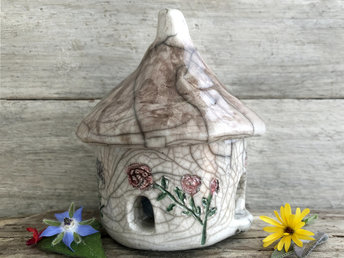 Round raku ceramic kurinuki cottage glazed in white with flowering plants on the walls in red-pink, purple, and blue. The roof has a chimney and is glazed to look like thatch. This house has a cute little doorway and two windows.