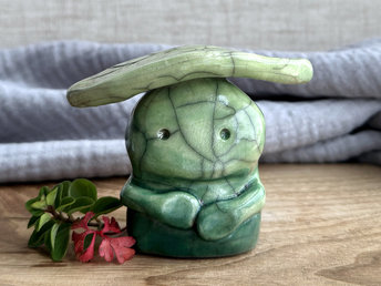 Raku ceramic sculpture glazed in forest green to spring green (bottom to top) with a kind, but slightly intense, face, and a brighter yellow-green leaf on its head. It also has two cute little arms.