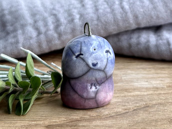 a smiley and loving Jizo Shinto raku ceramic sculpture talisman glazed in a bottom-to-top gradient of purple to blue. they have a kind, gentle face, and there's a small metal loop coming out of the top of their head that would allow them to be hung from something.