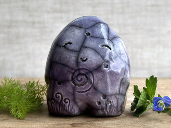round and vaguely egg-shaped nature spirit sculpture. it is glazed in a bottom-to-top gradient of purple to indigo violet and has spiral carvings on its body. it also has a gentle, happy face.